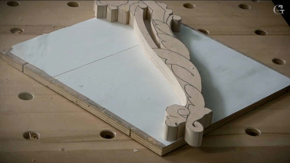 A shaped molding blank clamped to the bench beside its paper pattern, profile run but no ornament carved yet