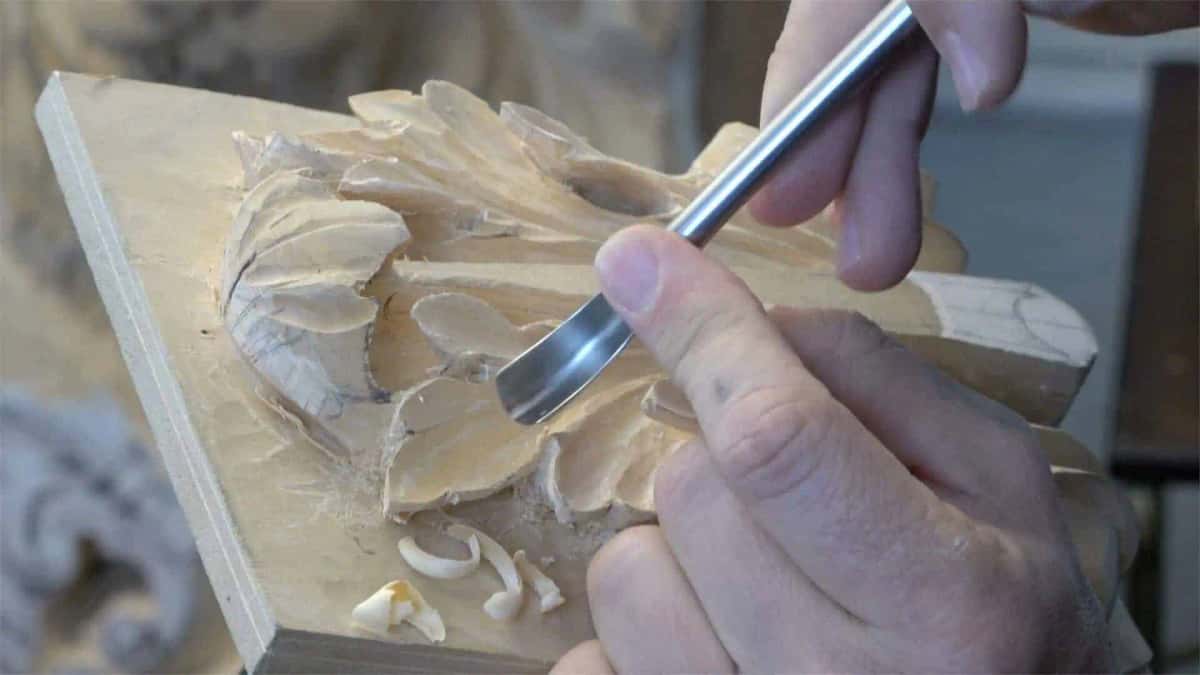 Fingers braced against a bright gouge blade cutting the lobes of a pale acanthus leaf