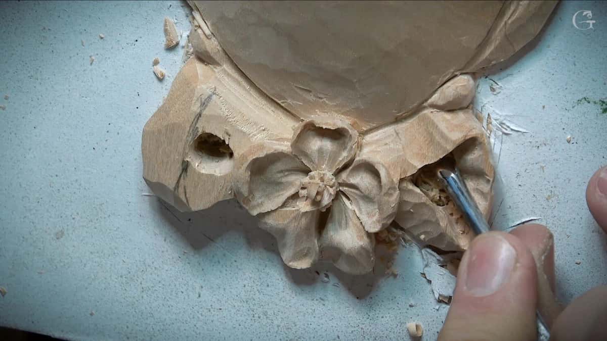 Close view of a gouge cutting in under the petals of a carved pale flower rosette, chips lifting away
