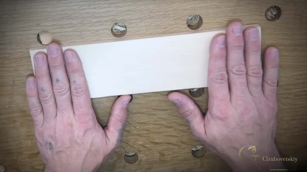 Two hands hold a pale basswood plank flat on a ray-flecked oak bench top between the bench dogs.