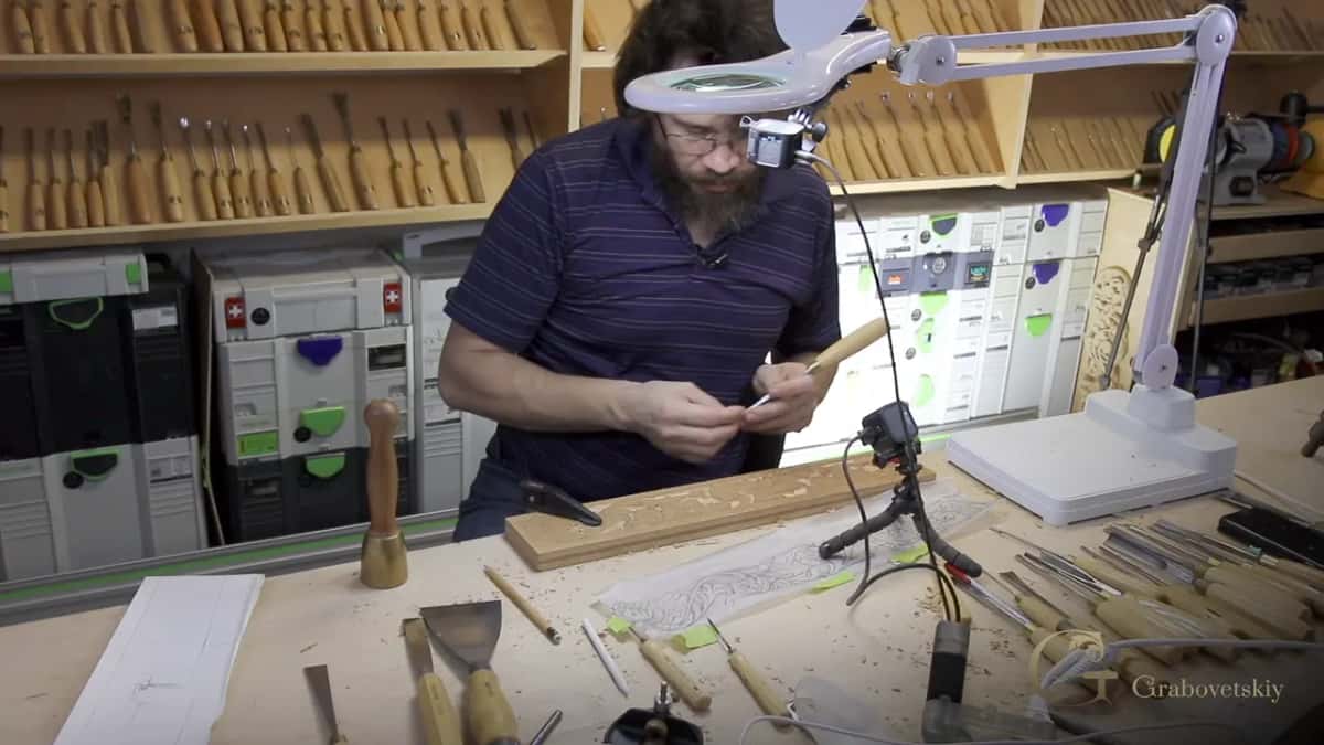 Alexander at his bench under a magnifier lamp, a long tan board and its paper pattern below a wall of gouges.