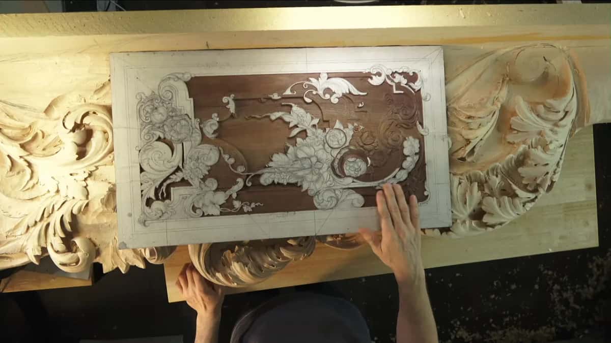 Hands hold a dark hardwood panel whose carved flowers are chalked white, above a pale carved frieze on the bench.