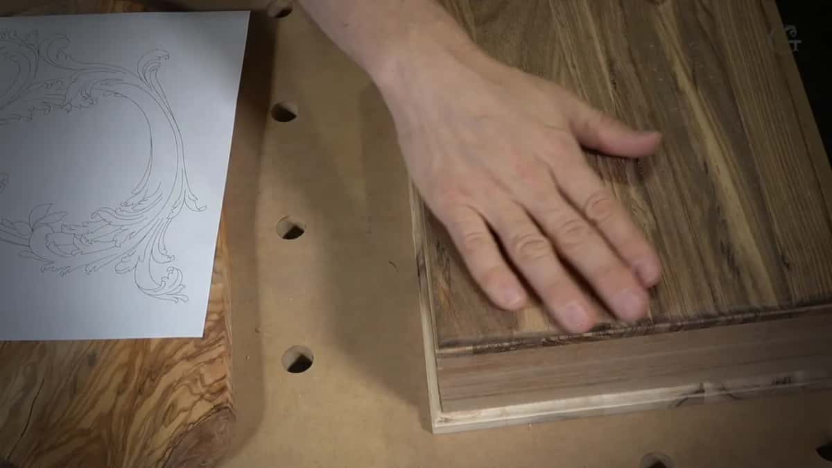 A hand rests flat on a glued panel of dark-streaked hardwood, a wildly figured board and a printed design beside it.