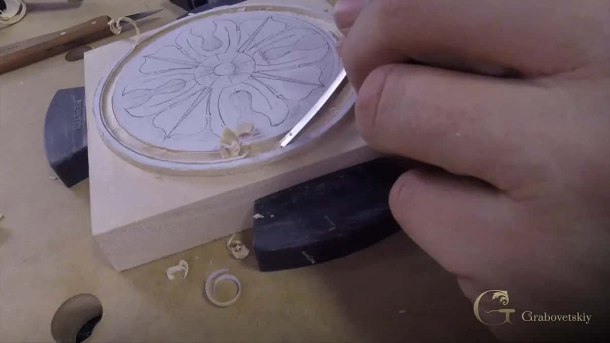 A round blank of pale wood with a rosette drawn on in pencil, a skew chisel starting the outline, mallet in hand.