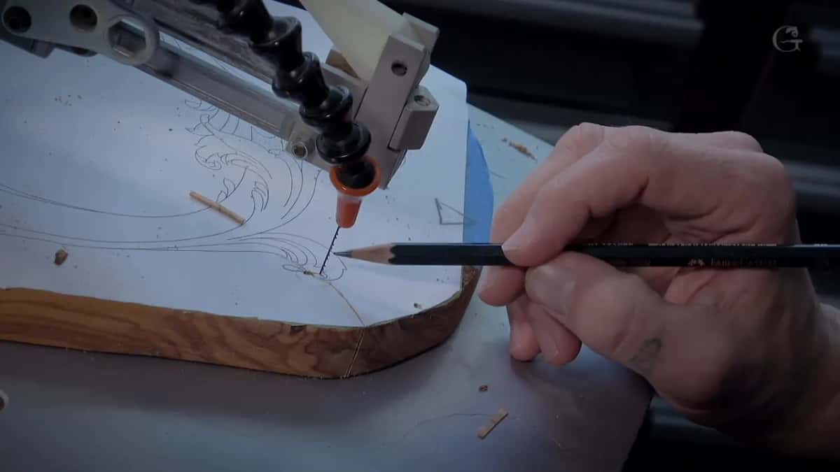 A scroll saw blade follows a line on a paper-covered blank; the sawn edge below shows gold wood with dark wavy streaks.