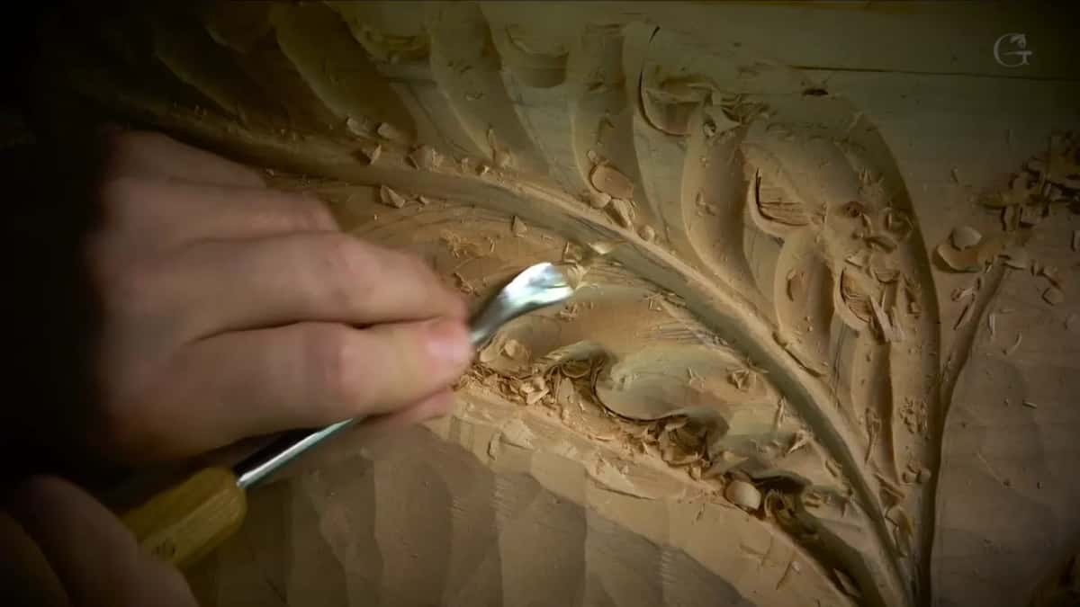 A bent shank with a yellow handle works close to the carved edge while the other fingers guide it