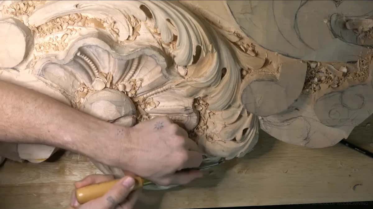 An orange-handled gouge, held low, carving into the lobe beneath the beaded shell while chips pile up around it.