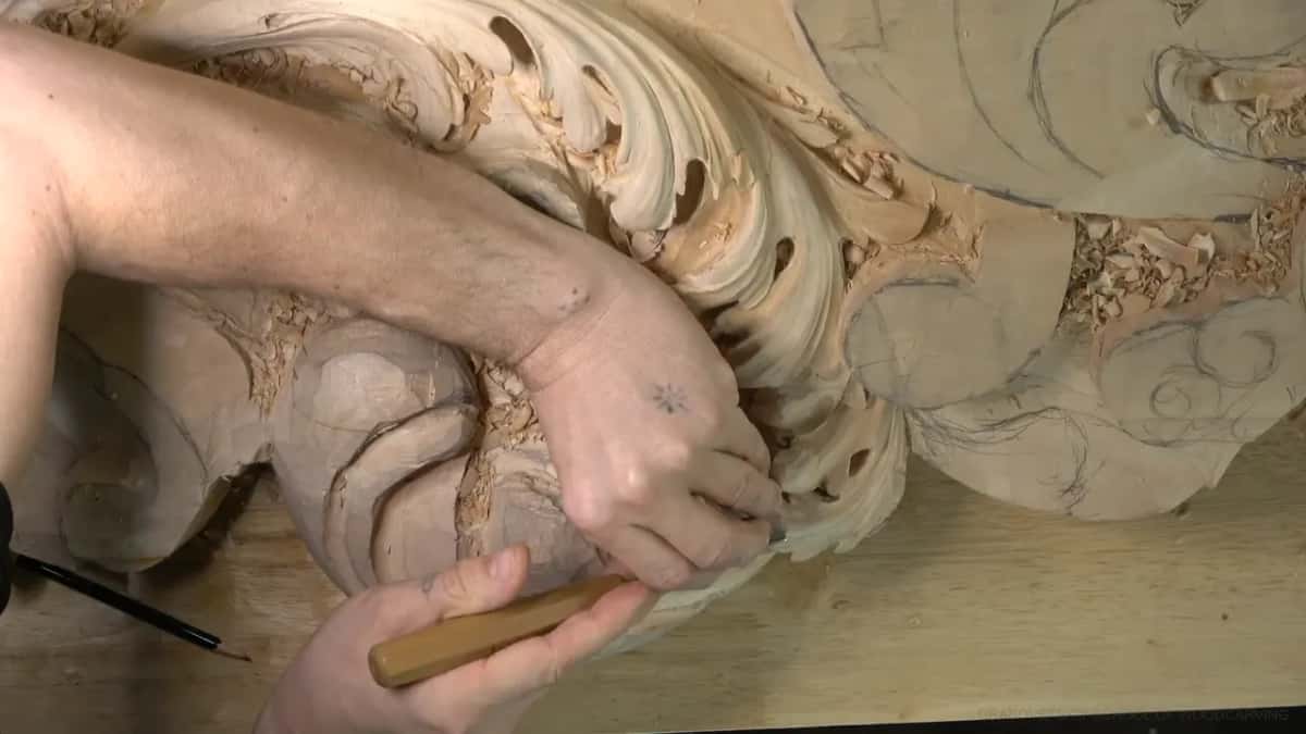 Arm across the panel, gripping a long yellow gouge at the far left curl, pencil on the bench.
