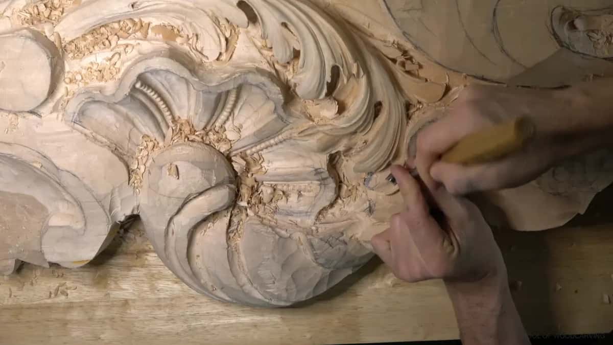 A yellow handled gouge pressed into an acanthus lobe beside the carved shell, both hands on the tool.