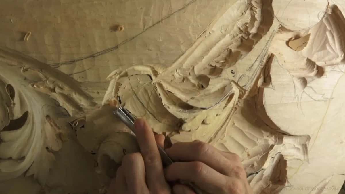 His right hand steers a gouge along a penciled edge while the curled scroll sits below it