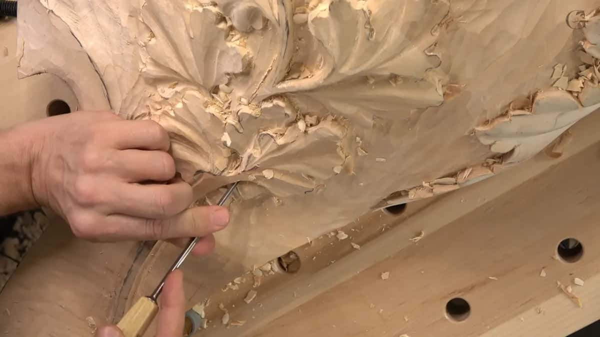 Farther back now, a hand works a thin gouge where the lowest leaves meet the stem, chips on the bench