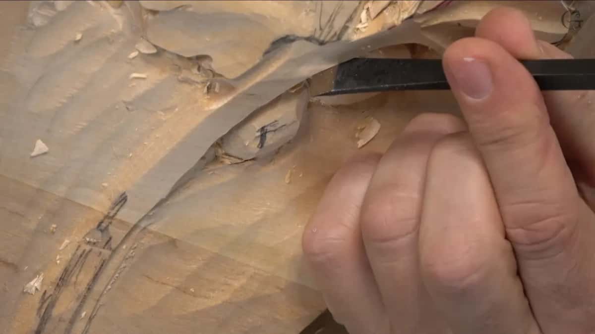 Fingers pinch a dark handled gouge above two rough lumps, still boxy and marked with pencil