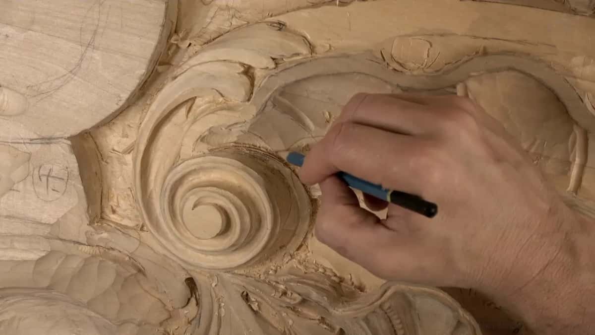A blue pencil runs along the edge of the carved spiral, the fingers of that hand resting on the wood.