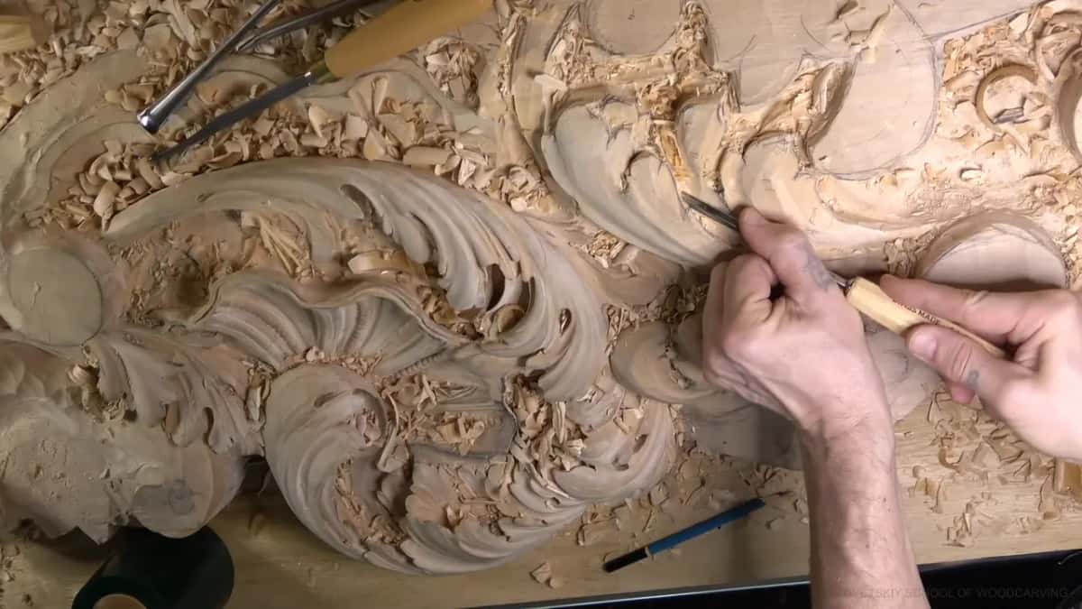 Wide shot, both hands carving at right, the big finished head at left.
