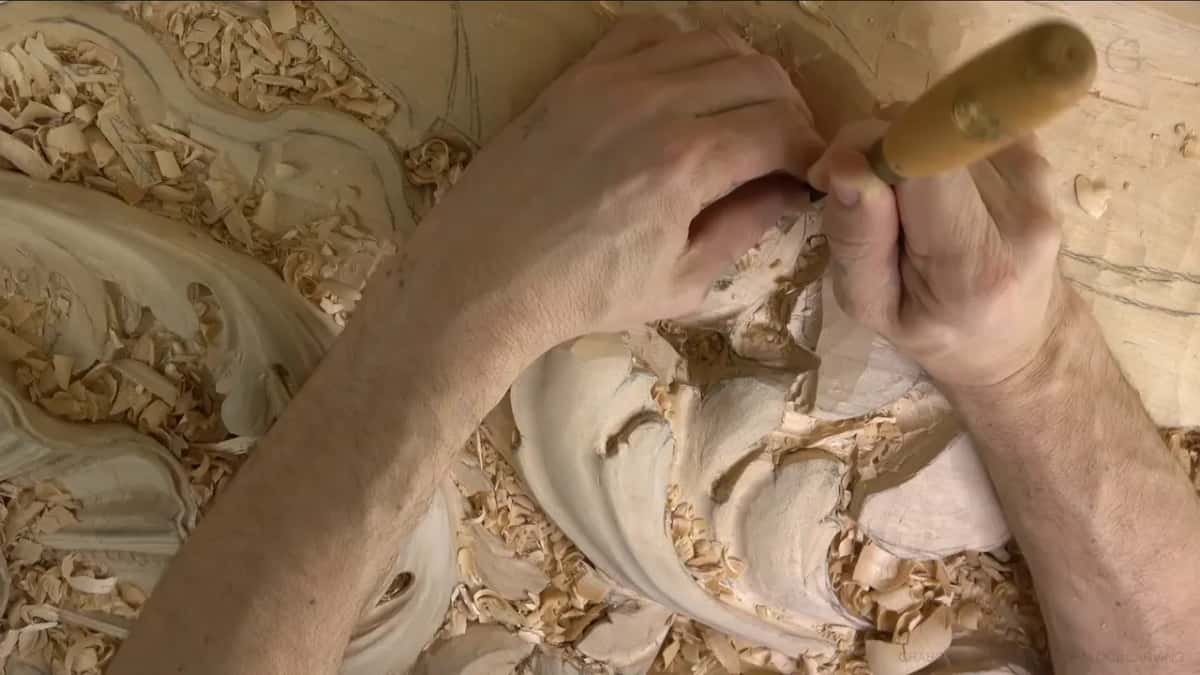 Both hands guide one gouge, the handle tipped up and the blade working low in the leaf beside a curl.