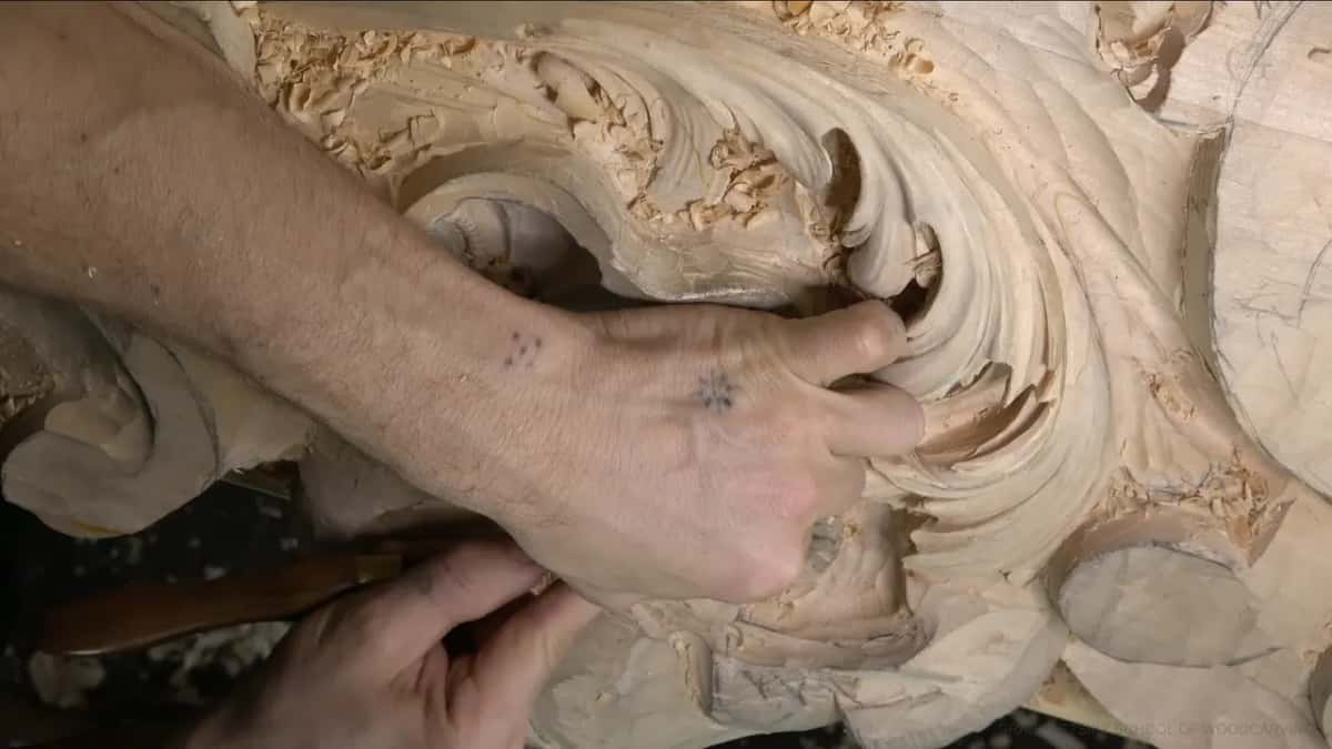 One hand presses a small gouge into a curled lobe while the other steadies a dark handle.