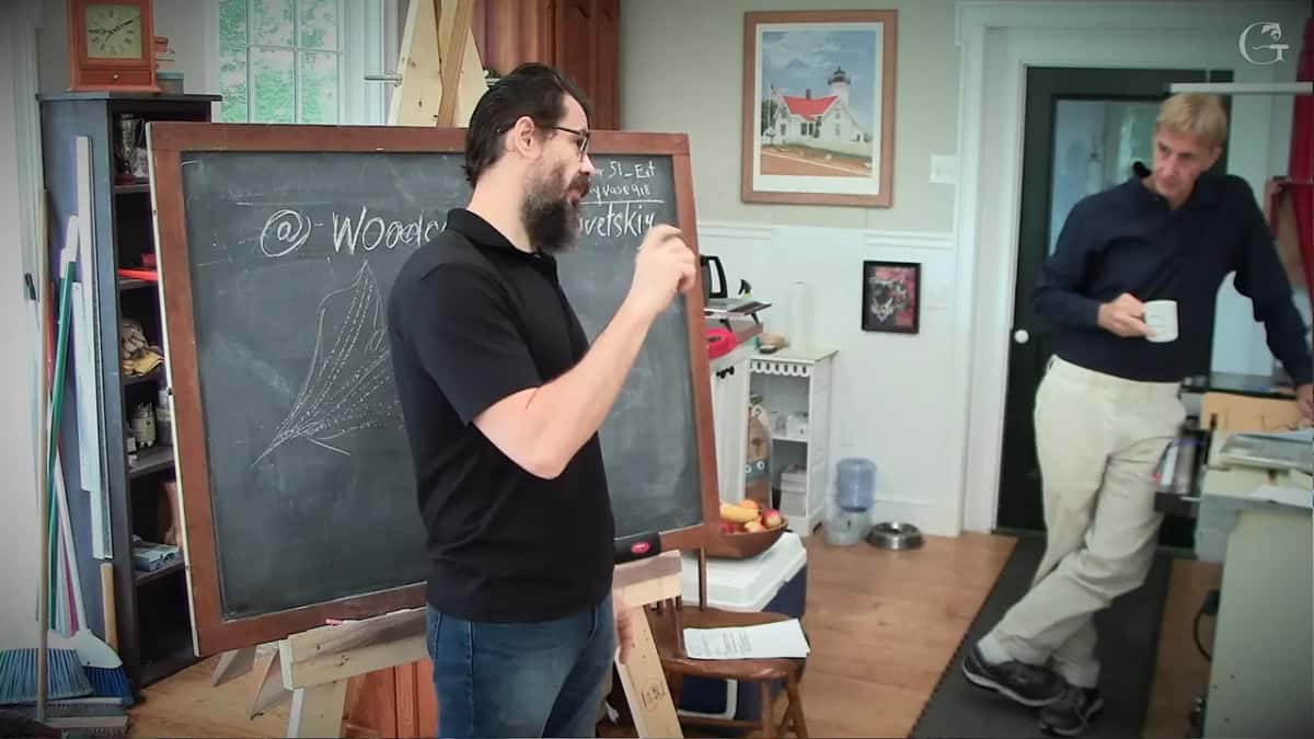 At the chalkboard with a finger raised, a student holding a mug of coffee and waiting
