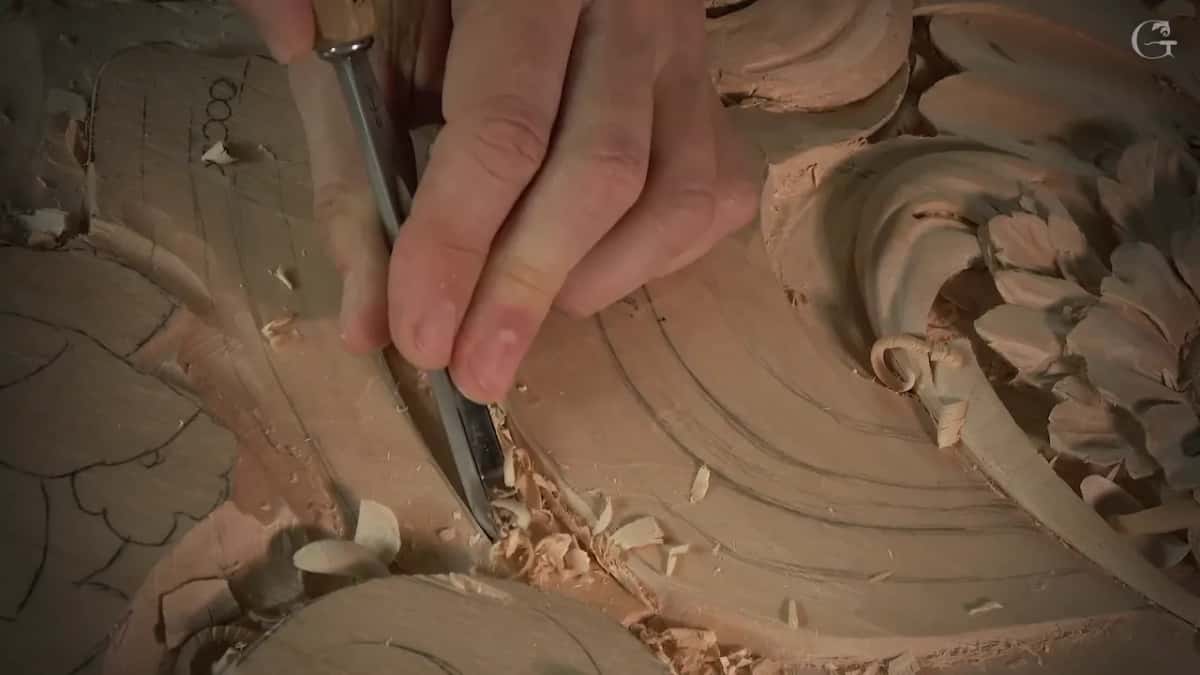 Fingers steady a gouge at the rim of the groove next to the big scroll