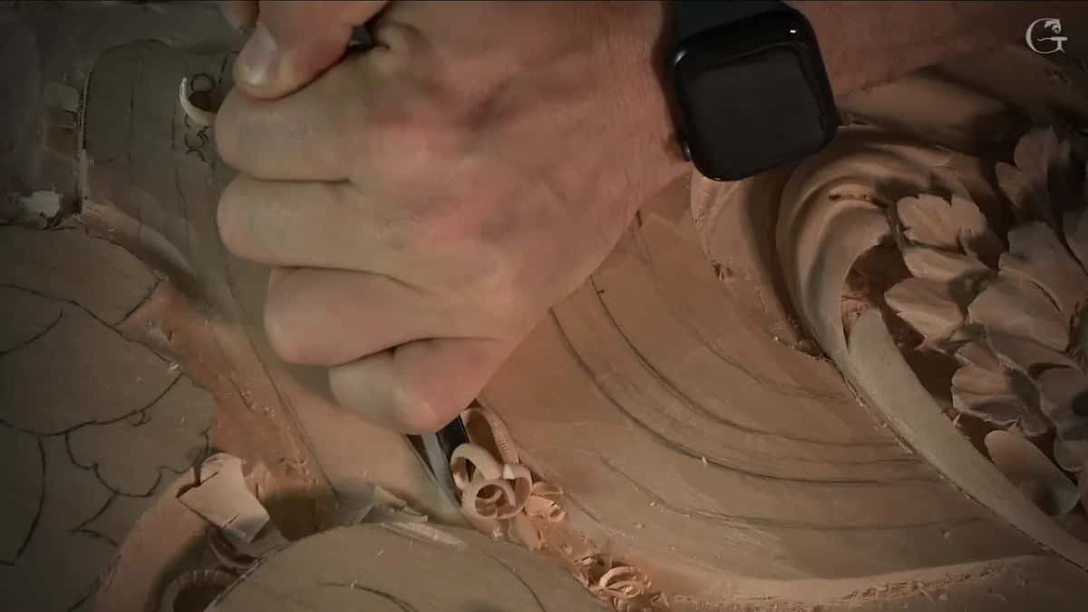 A hand grips the gouge low while its tip rides a groove, curls of wood piled beside it