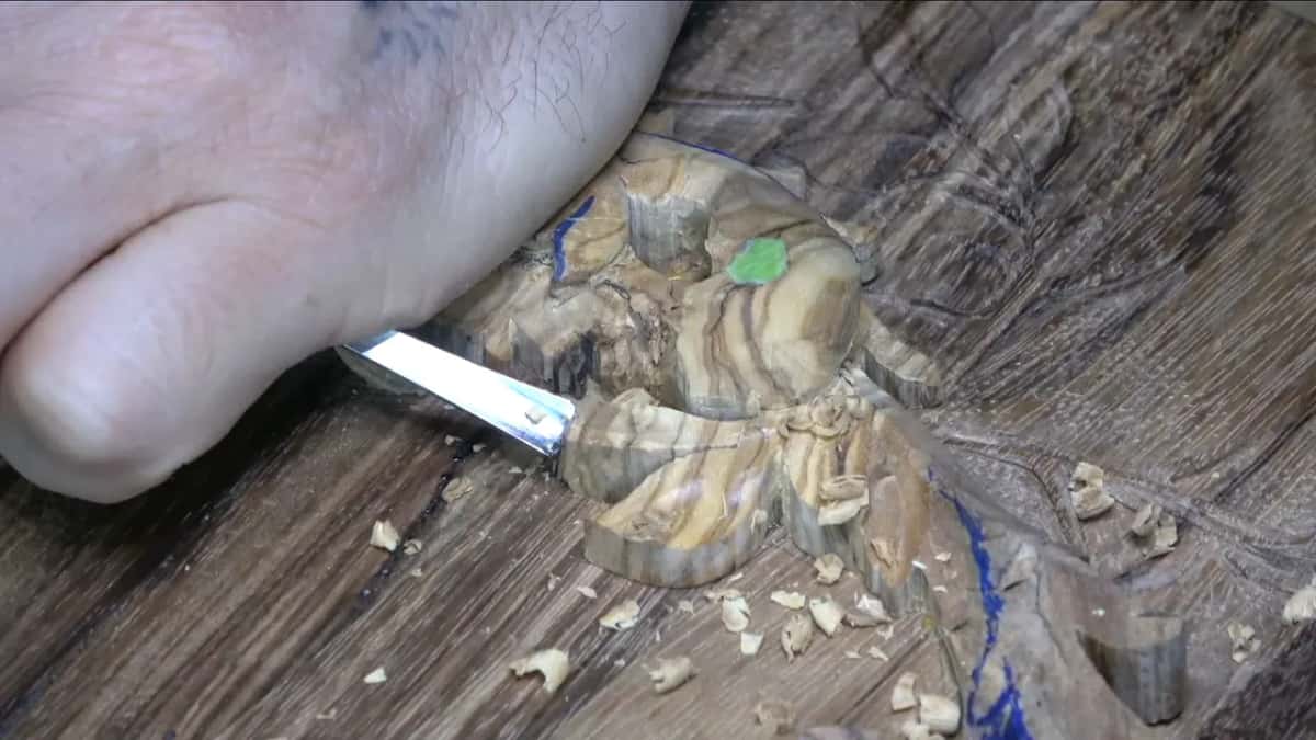 Flat blade under a pressing hand lowers a streaky olive lobe toward the lid