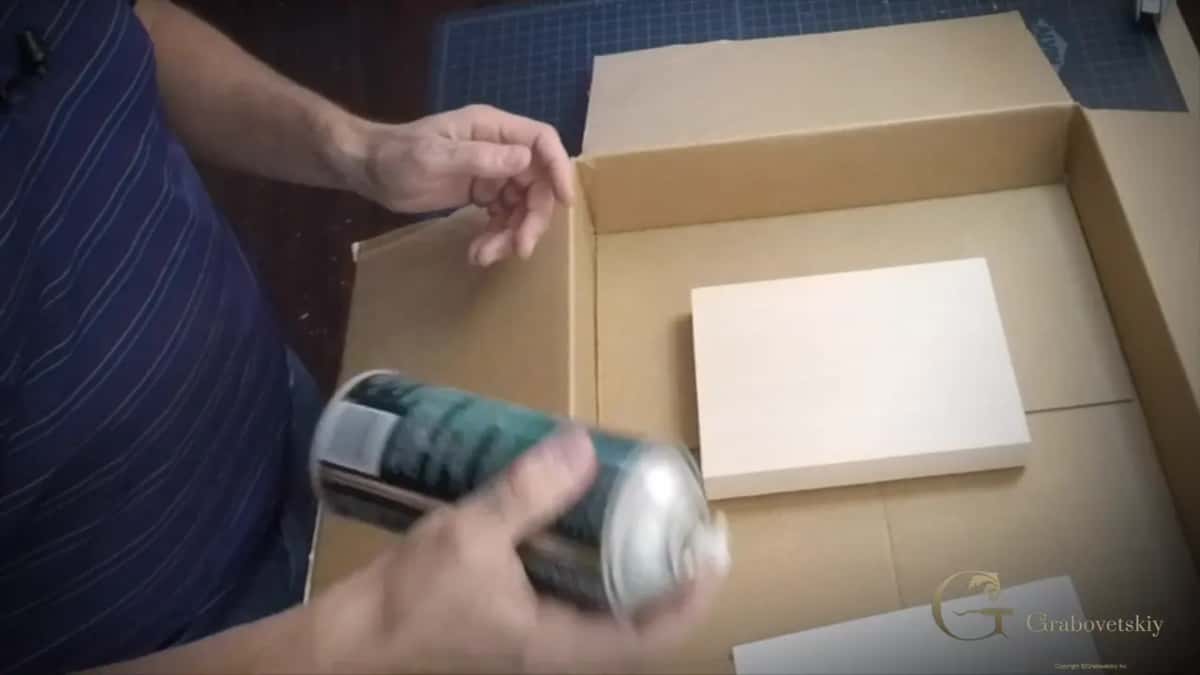 The spray can angled over the block inside a cardboard box, which keeps the glue off everything else