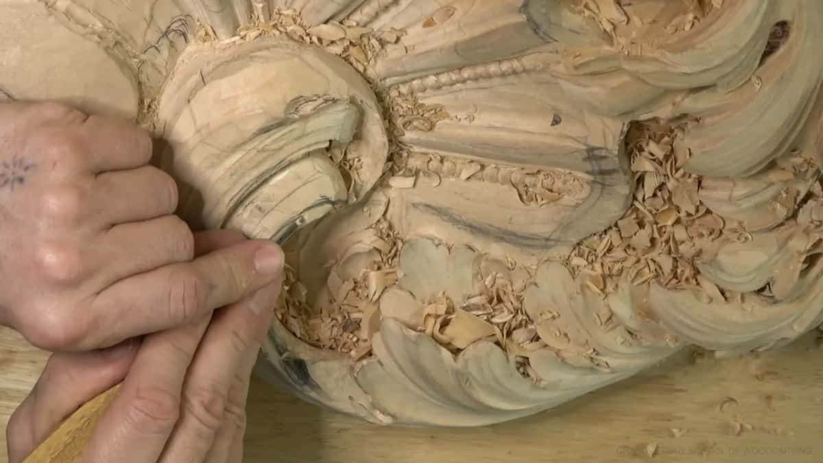 A hand grips a gouge inside the scroll while chips pile up on the leaves below.