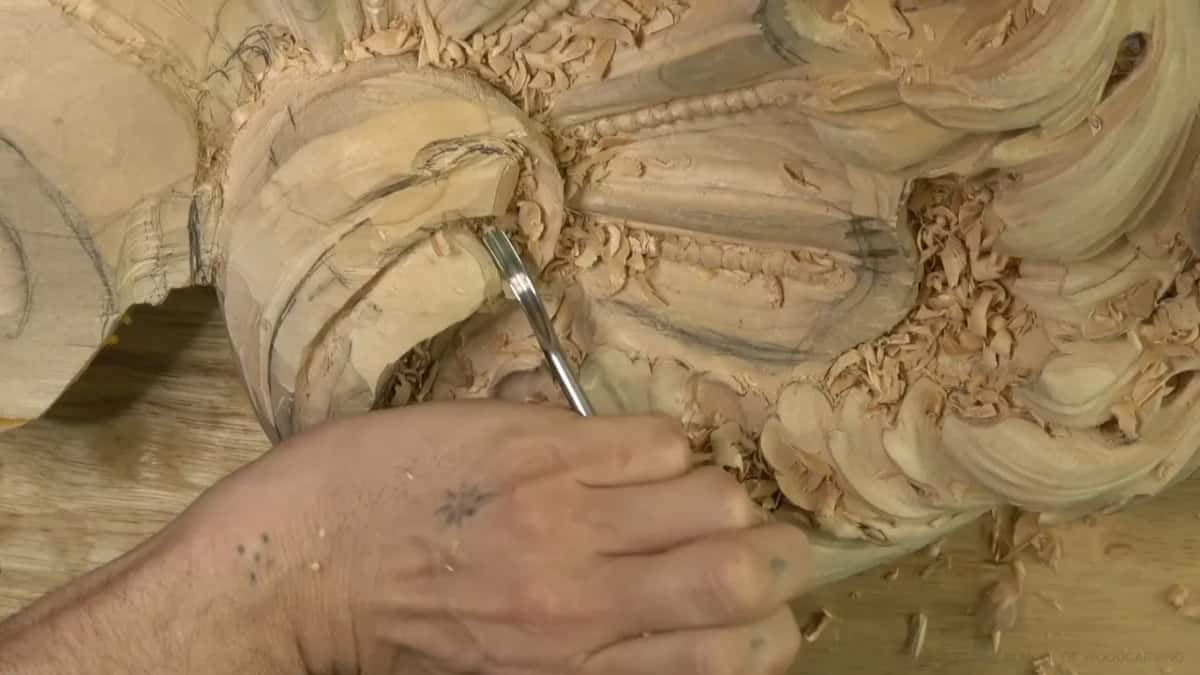 Fingers pinch a slim gouge deep in the hollow of the scroll, with loose chips resting on every leaf.