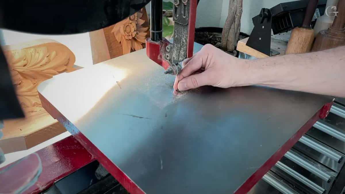 Hand pointing at the hair thin scroll saw blade, red machine, carved panels stacked behind on a shelf