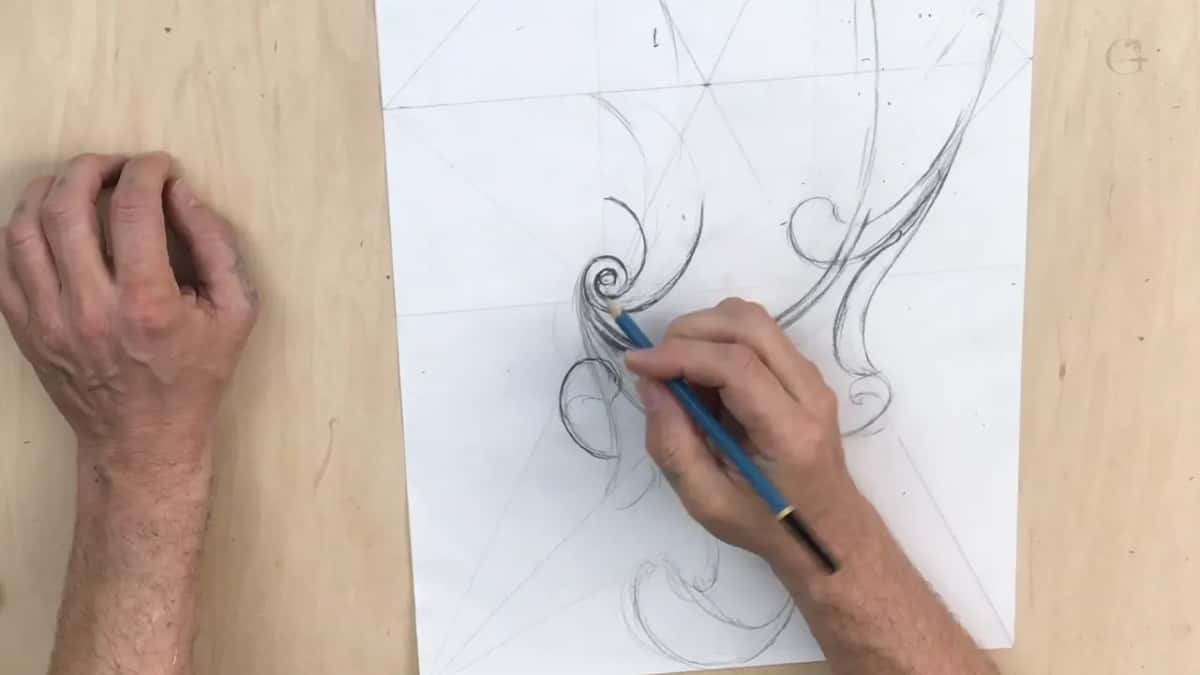 Blue pencil over the paper drawing, geometric construction lines ruled across the sheet before any scroll was drawn