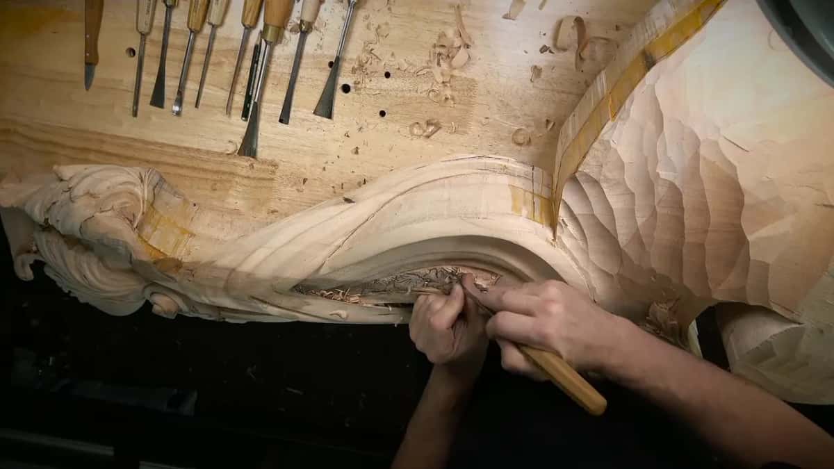 Gouges in a row on the bench while two hands carve inside the curved panel of the corbel side