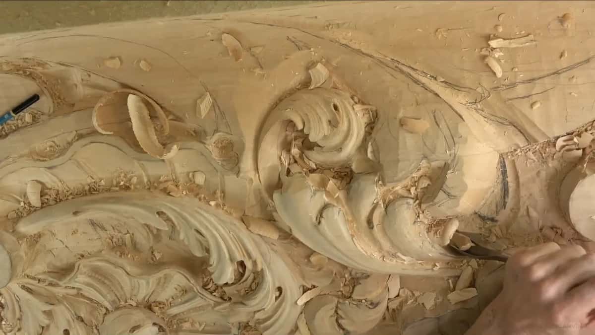 Deep acanthus scrolls, shavings everywhere, and a hand working a gouge into one hollow