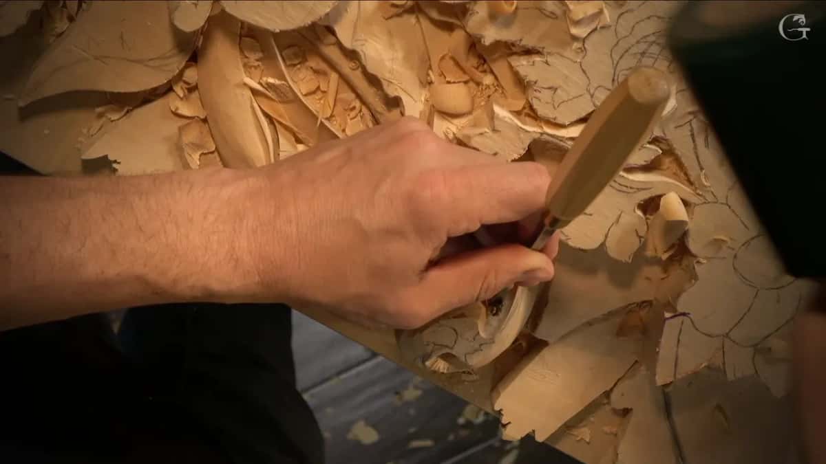 Hand pressing a gouge straight down into the carved scroll, loose shavings curling away on both sides of the blade