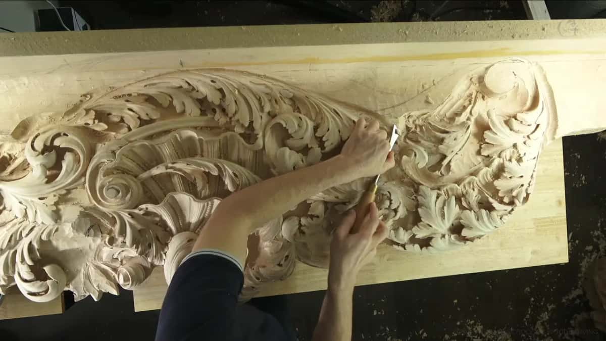 From above, the whole ceiling board with its acanthus scrolls, two hands working near the right end.