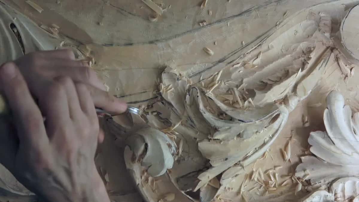 Fresh chips pile up while a gouge follows a pencil line above the acanthus.
