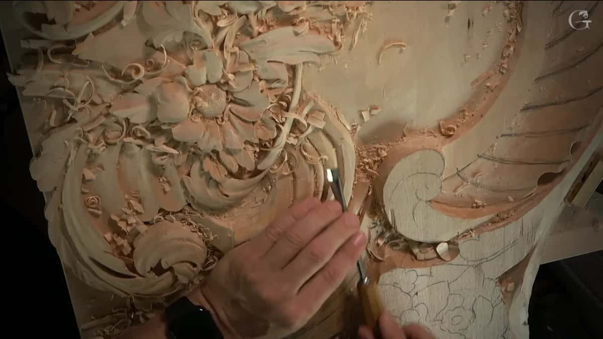 Two hands hold a gouge beside the carved flower, a small scroll just to its right