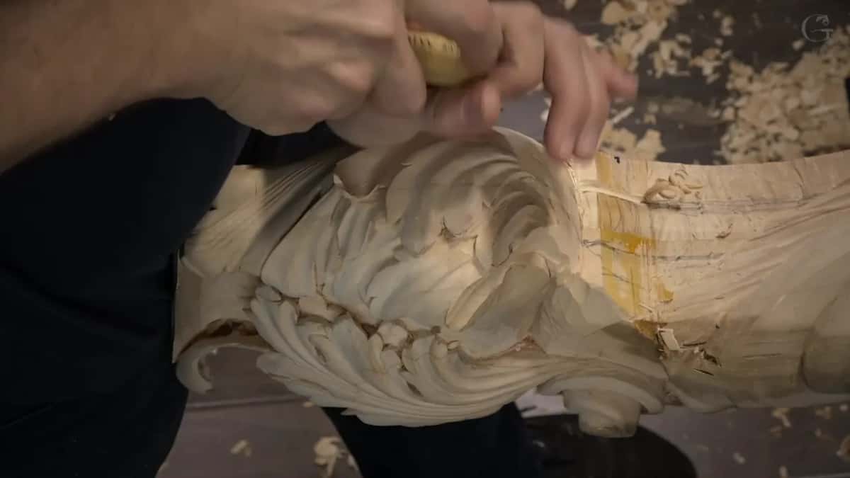 Two hands drive a gouge where the acanthus meets the yellow-marked middle.