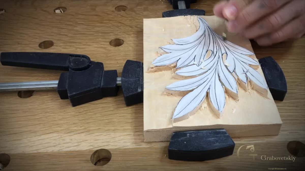 Block clamped at an angle, and one hand bringing a light handled gouge down to the upper edge of the carving.