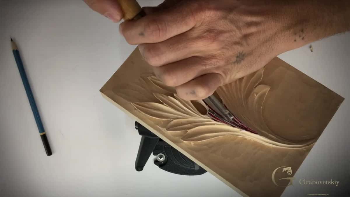 A light handled gouge gripped low, cutting toward the middle of the leaf, with the wood lifting ahead of it.