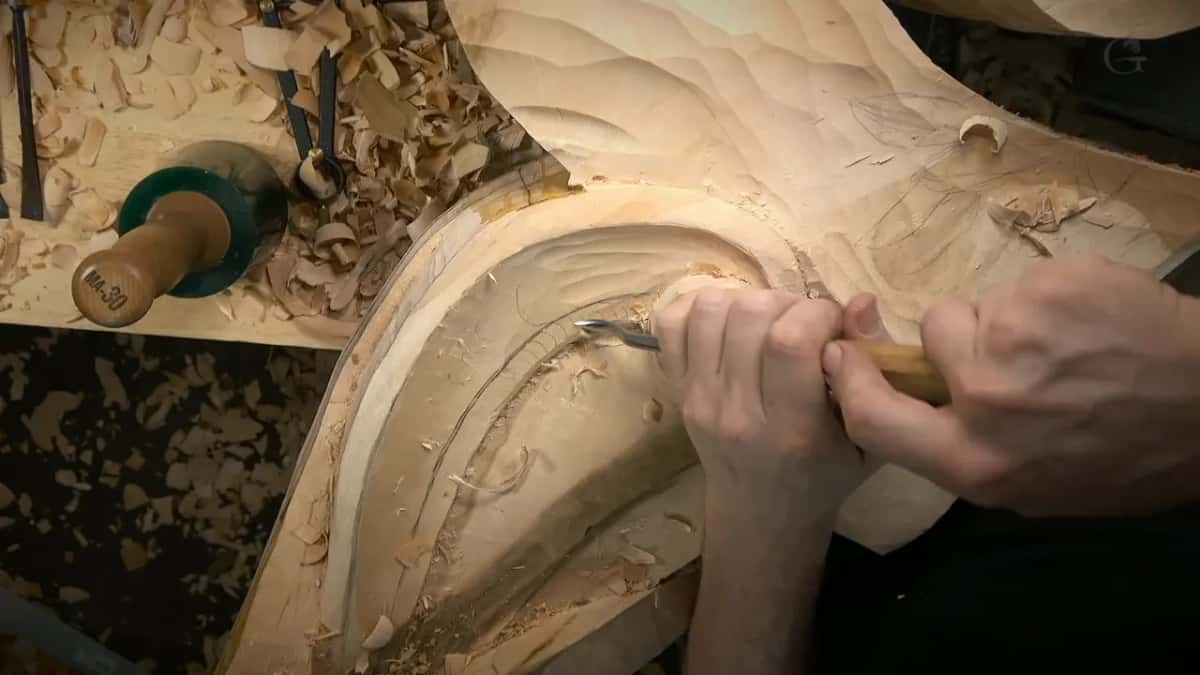 Fists around a pale handle push a curved gouge along the rounded side, a mallet standing at upper left