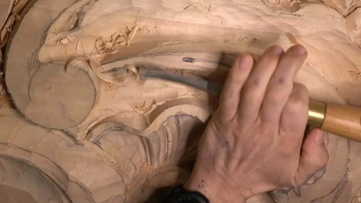 A wide gouge with a brass ferrule carves the long slope of the leaf, following a pencil line.