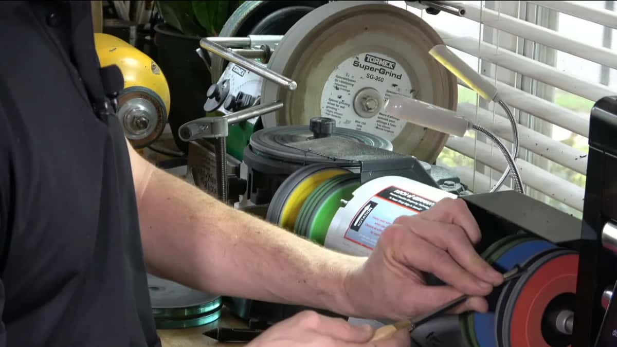 Both hands charge a stack of red and blue wheels with a compound stick, the Tormek and a yellow buffer behind