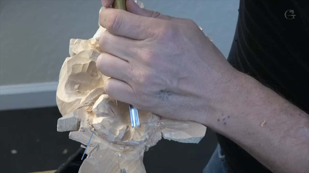 Fingers spread across the top of the flower head to hold it while a narrow tool works the hollow below, grey wall behind