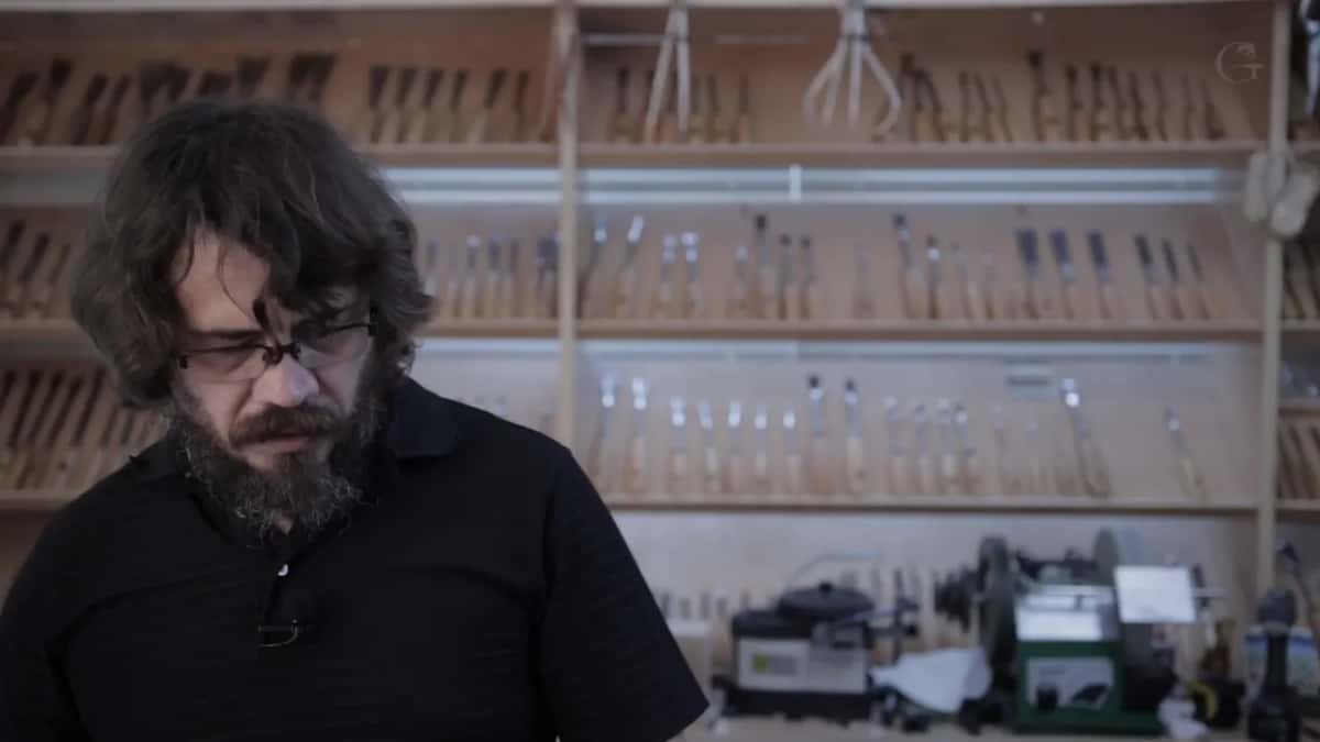 Alexander at the bench with four shelves of gouges and chisels out of focus on the wall behind him