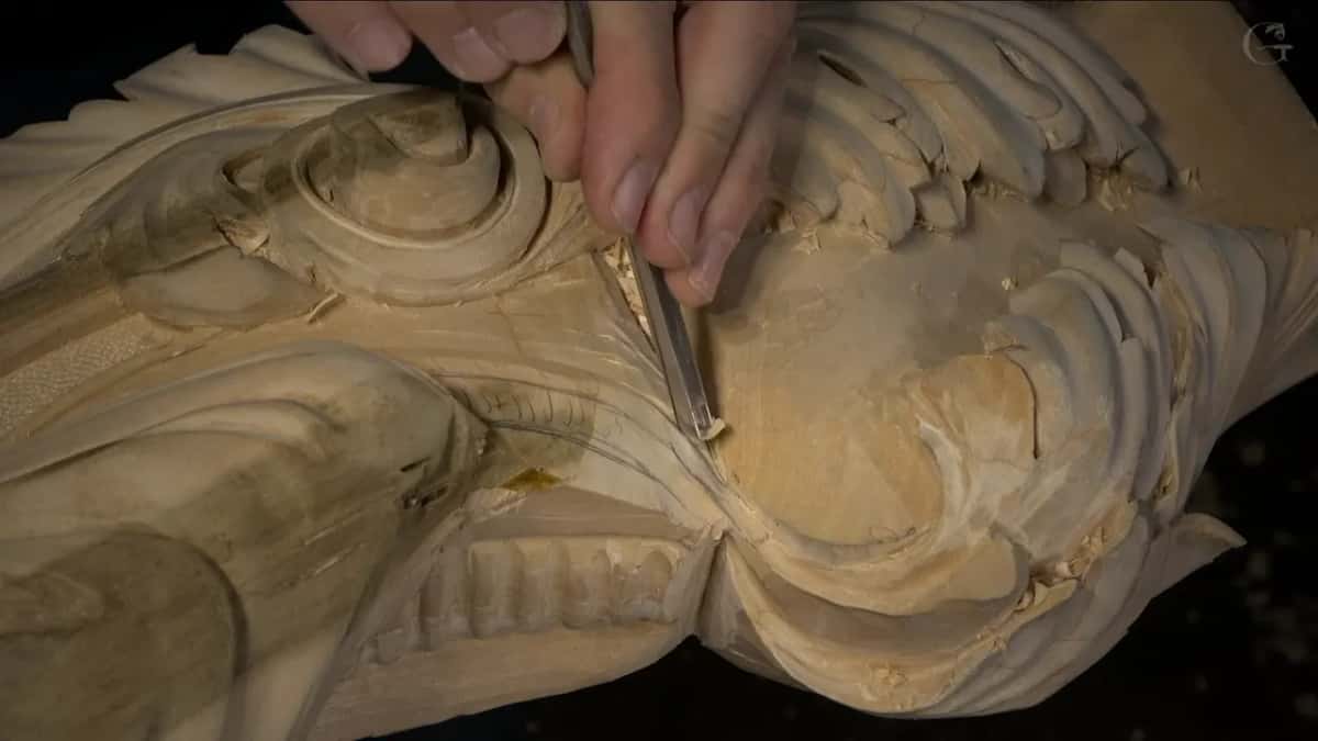 Straight gouge guided by two fingers along the side of a carved spiral, with a ribbed band running below it.