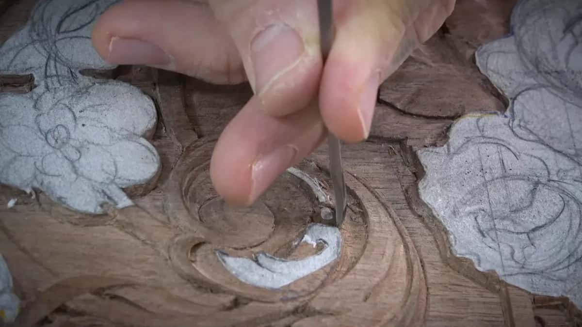 Slender blade held vertically between two fingers above the carved scroll, shavings around it
