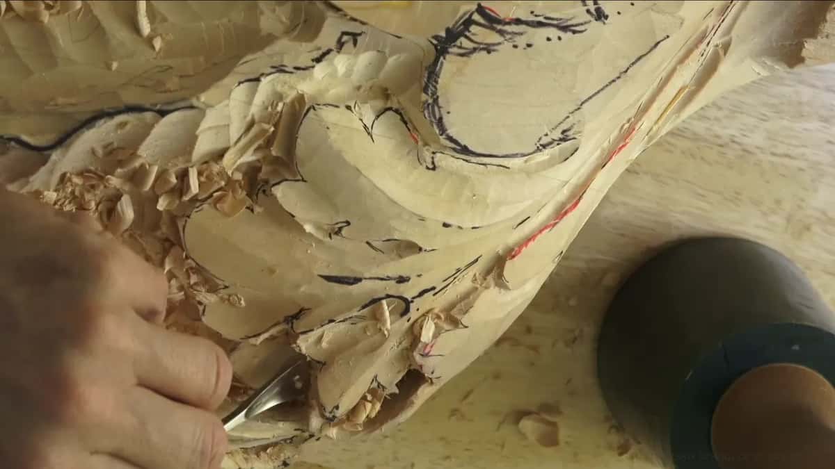 Fingers on a gouge in the carving, a green mallet resting on the bench beside it.
