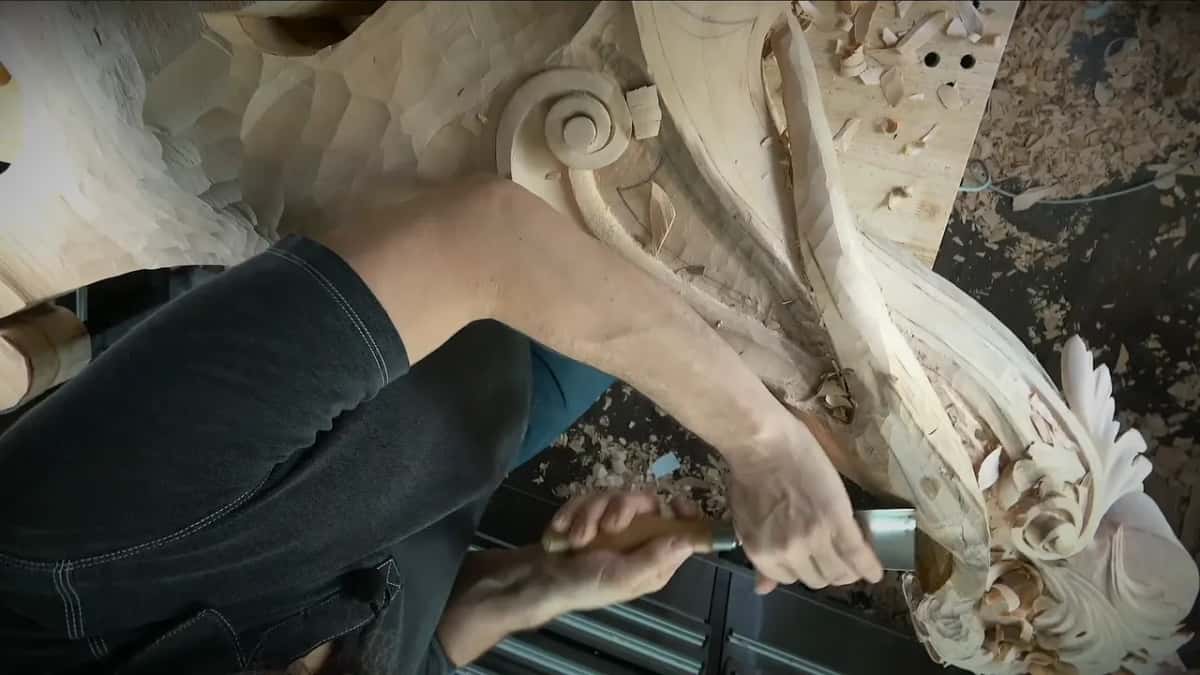 Right hand pushing a gouge where the twisted band meets the leaves, left arm across the bench