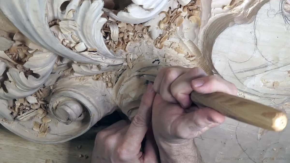 Two hands steer a long-handled gouge into the hollow low on the rolled head, shavings piled above.