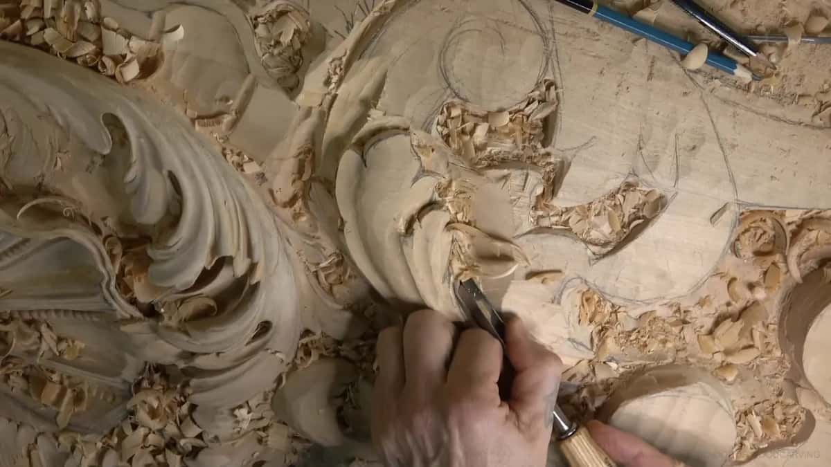 Fingers guide a pale-handled gouge into one curl of the new leaf.