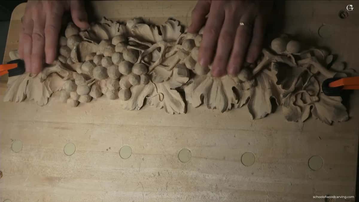 Two hands resting on the carved grapes and leaves, orange clamps holding each end of the board